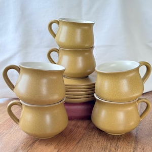 May include: A white ceramic mug with a black handle and the text "But First, Coffee" in a bold, black font. The mug is set against a blurred background, highlighting its simple design.