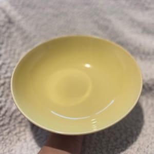 May include: A pale yellow ceramic bowl with a rounded shape. The bowl has a smooth, glossy finish and appears to be empty. The bowl is held by a person's arm. The bowl is likely for serving food.