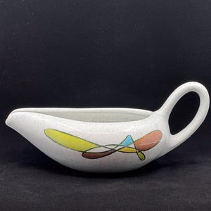 May include: A white ceramic gravy boat with a speckled finish and a curved handle. The exterior features a colorful abstract graphic design consisting of intersecting shapes in yellow, brown, light blue, and peach tones along the side.
