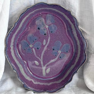 May include: A handmade ceramic plate with a scalloped edge. The plate features a vibrant purple glaze with a floral design in the center. The flowers are a lighter shade of purple with dark blue accents. The rim has a silver-colored trim.