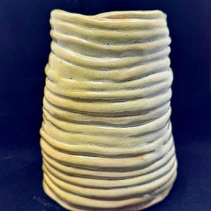 May include: A cylindrical ceramic vase with a glossy, light yellow-green glaze. The exterior surface features a series of horizontal, raised, undulating ridges that wrap around the entire body, creating a textured, ribbed appearance throughout.