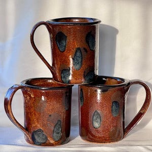 May include: A white ceramic mug with a black handle and the text "But First, Coffee" in a black, bold font. The mug is sitting on a wooden surface. The mug is approximately 4 inches tall.