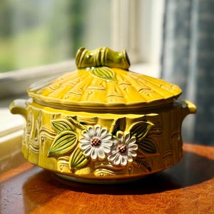 May include: A round yellow ceramic casserole dish with a matching lid, featuring a raised bamboo-like texture and a decorative relief of two white daisies with green leaves on the front. The lid has a central handle shaped like bamboo stalks.