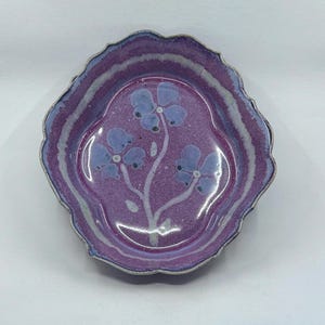 May include: A scalloped ceramic dish with a glossy purple glaze and a hand-painted light blue floral motif in the center. The edges of the dish feature a lighter purple and white gradient, creating a distinct border around the central flower design.