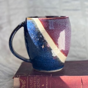 May include: A handmade ceramic mug with a unique design. The mug features a dark blue base, transitioning to purple and cream diagonal stripes. It has a black handle and is set on a vintage book.