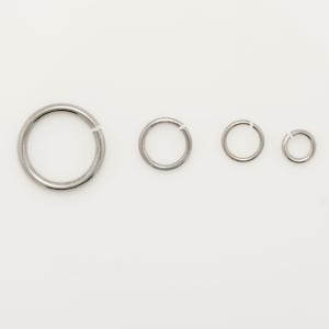 May include: Four silver-colored jump rings of varying sizes are arranged on a white background. The rings are open, with a small gap in the circle. The sizes range from large to small, suitable for jewelry making or crafts.