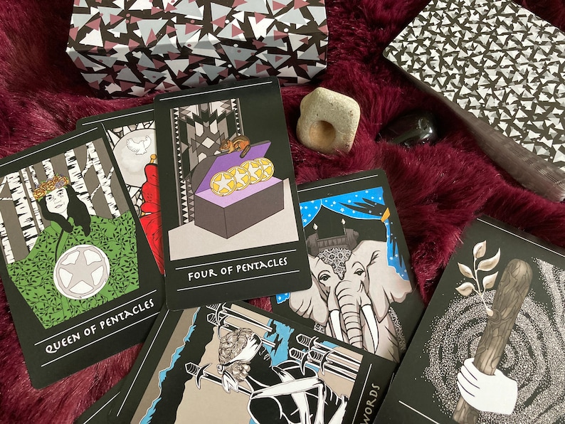 May include: A tarot card reading spread with ten cards laid out on a burgundy fuzzy surface. The cards are black with white and colored illustrations. The cards include "Queen of Pentacles", "Four of Pentacles", and "Three of Swords".
