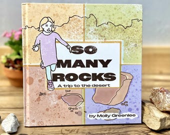 May include: A square hardcover book titled So Many Rocks: A trip to the desert by Molly Greenlee. The cover features a cartoon illustration of a child in a light purple dress, set against a background of four colored panels with rock graphics.