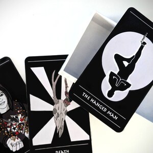 May include: Three tarot cards with black backgrounds and white illustrations. The card on the left shows a woman with dark hair and a black and white patterned shirt. The card in the middle shows a white skull with antlers and flowers. The card on the right shows a silhouette of a person hanging upside down with the text "The Hanged Man".
