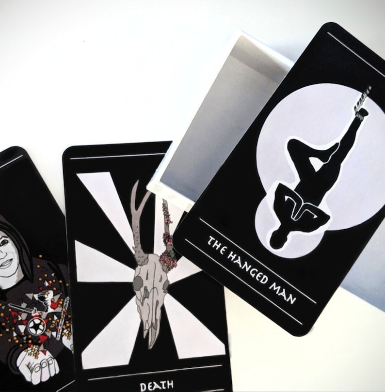 May include: Three tarot cards with black backgrounds and white illustrations. The card on the left shows a woman with dark hair and a black and white patterned shirt. The card in the middle shows a white skull with antlers and flowers. The card on the right shows a silhouette of a person hanging upside down with the text "The Hanged Man".