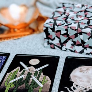 May include: Three tarot cards laid out on a gray surface. The card on the left features a man with a turban and a key symbol. The middle card shows three swords crossed in a desert landscape. The card on the right shows a scorpion with a full moon in the background.