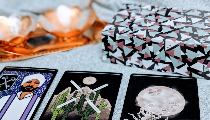 May include: Three tarot cards laid out on a gray surface. The card on the left features a man with a turban and a key symbol. The middle card shows three swords crossed in a desert landscape. The card on the right shows a scorpion with a full moon in the background.
