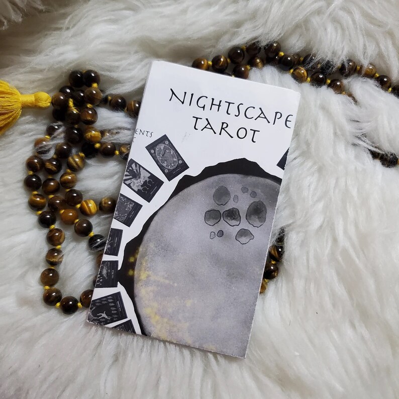 May include: A white book with the title "Nightscape Tarot" printed in black. The book is lying on a white fluffy surface with a brown and gold beaded necklace.