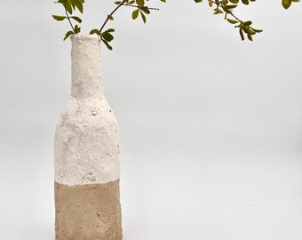 May include: A white and brown ceramic vase with a rough texture, holding a sprig of green leaves.