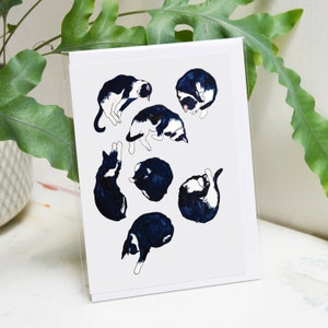 May include: A white card with a black and white illustration of seven cats in various sleeping positions. The cats are drawn in a simple, stylized manner.