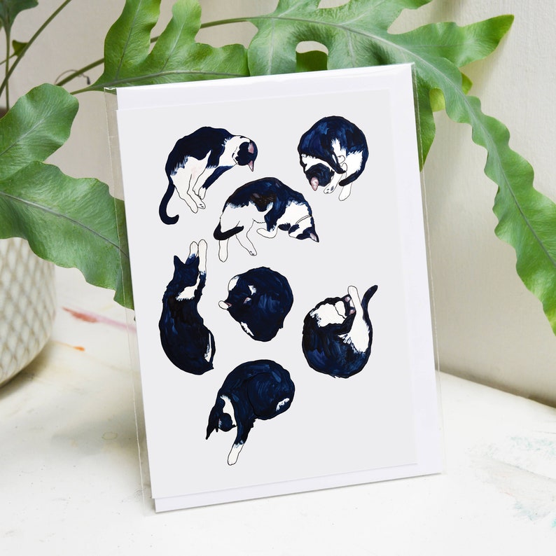 May include: A white card with a black and white illustration of seven cats in various sleeping positions. The cats are drawn in a simple, stylized manner.