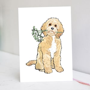 Könnte beinhalten: Eine Aquarell-Illustration eines hellbraunen Hundes mit weißen Abzeichen, der sitzt und einen Zweig Mistel in seinem Maul hält.