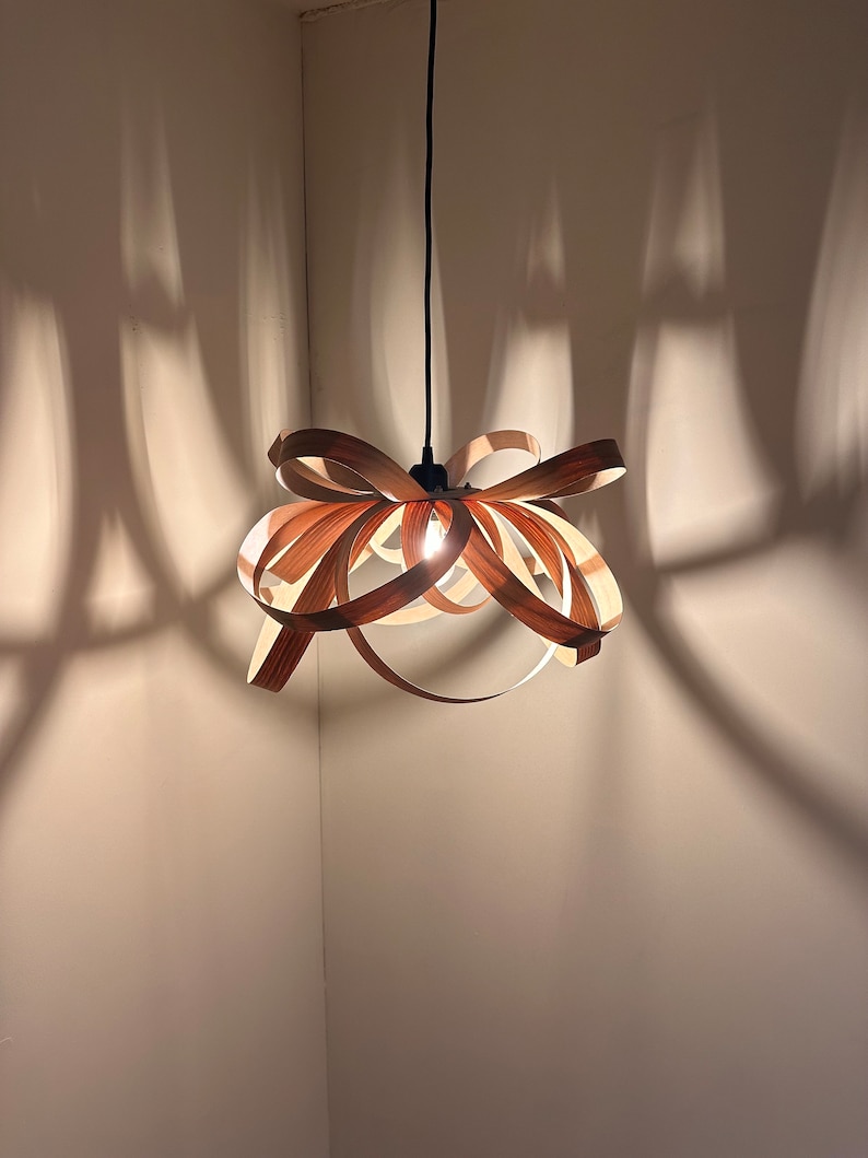 May include: A wooden pendant light fitting with a unique, interwoven design. The light fitting is hanging from a black cord and casts a warm glow.
