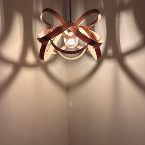 May include: A wooden pendant light fitting with a unique, interwoven design. The light fitting is hanging from the ceiling and casting shadows on the wall.