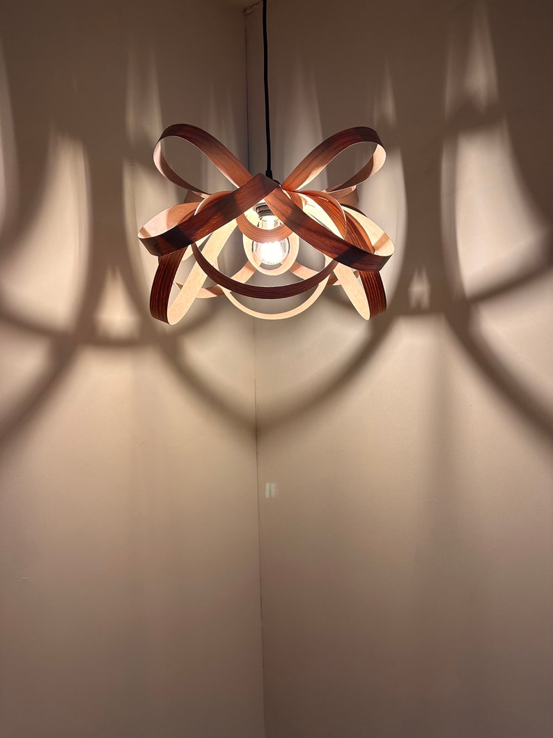 May include: A wooden pendant light fitting with a unique, interwoven design. The light fitting is hanging from the ceiling and casting shadows on the wall.