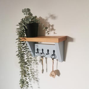 May include: A light blue wooden key rack with four hooks and a small shelf. The rack has a natural wood top and is mounted on a white wall. A potted plant hangs down from the shelf. The text "Our Home" is written on a small wooden sign hanging from one of the hooks.