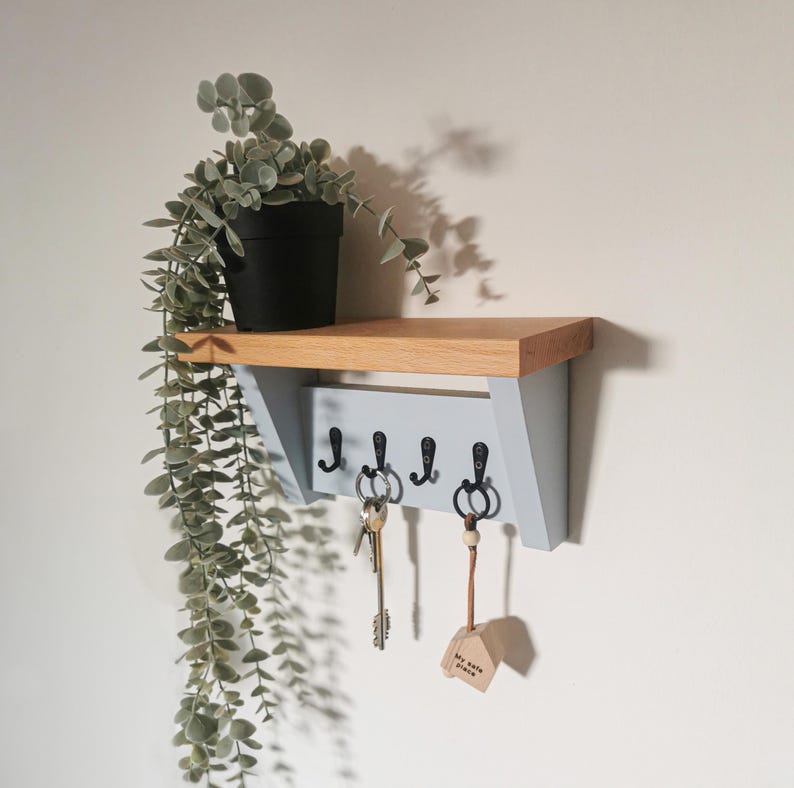 May include: A light blue wooden key rack with four hooks and a small shelf. The rack has a natural wood top and is mounted on a white wall. A potted plant hangs down from the shelf. The text "Our Home" is written on a small wooden sign hanging from one of the hooks.