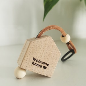 May include: A wooden house-shaped keyring with a brown leather strap and wooden beads. The house has the words "Welcome home" and a small heart engraved on it.