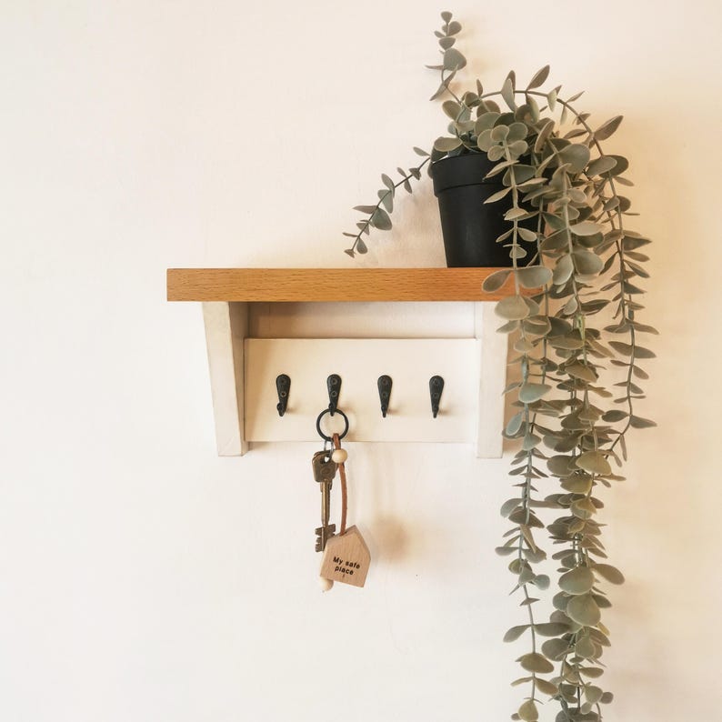 May include: White key rack with a wooden shelf and five hooks. A set of keys with a wooden house-shaped keychain that says "My safe place" is hanging on one of the hooks. A potted plant with trailing green leaves is on the shelf.