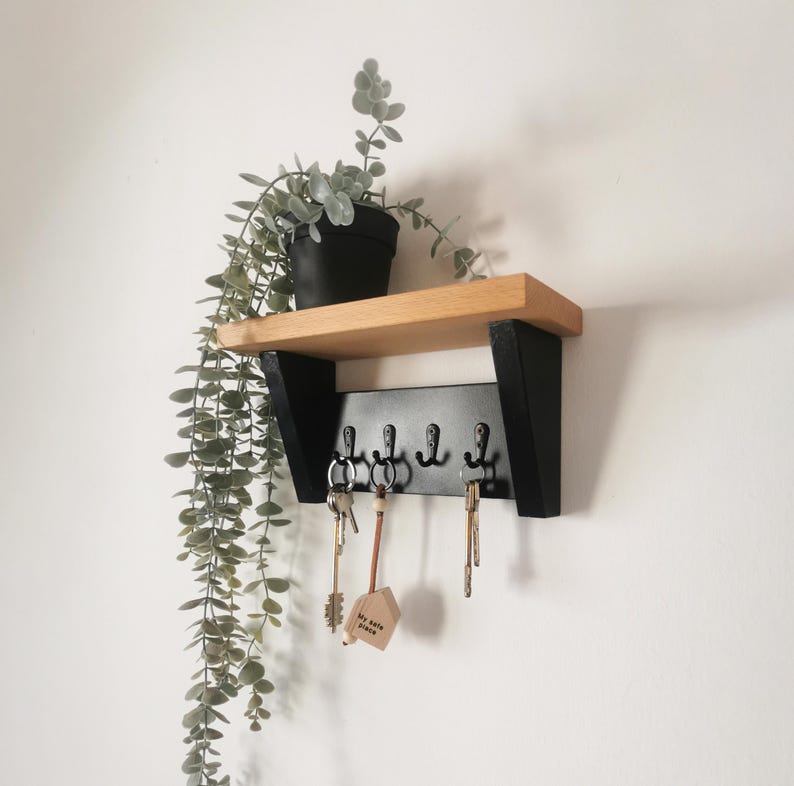 May include: A black key rack with five hooks mounted on a wooden shelf. The shelf is attached to a white wall. A potted plant with green leaves hangs down from the shelf. The key rack has a small wooden sign that says "My Home Place".