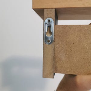 May include: Close-up of a wooden shelf with a metal bracket attached to the side. The bracket has a keyhole slot for hanging the shelf on a wall.