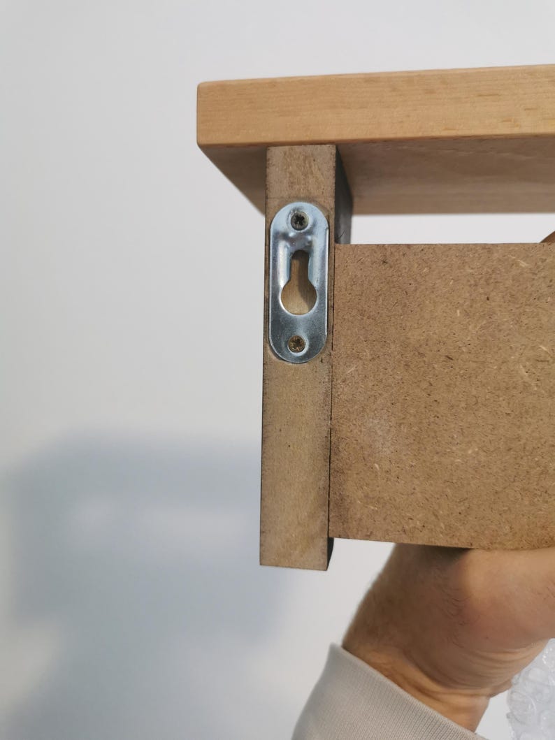May include: Close-up of a wooden shelf with a metal bracket attached to the side. The bracket has a keyhole slot for hanging the shelf on a wall.
