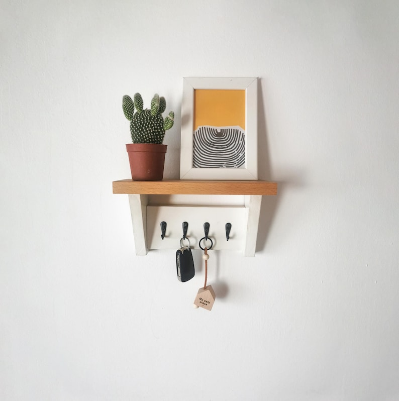 May include: White key rack with four hooks, a small potted cactus, and a framed print with a yellow and black geometric design. The key rack has a wooden shelf and is mounted on a white wall.