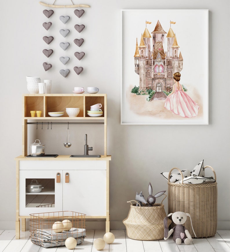 K&ouml;nnte beinhalten: Eine wei&szlig;e Spielk&uuml;che mit einem Holzrahmen und wei&szlig;en Schr&auml;nken. Die K&uuml;che hat ein Sp&uuml;lbecken, ein Kochfeld und ein Regal &uuml;ber dem Kochfeld. Auf dem Boden stehen zwei K&ouml;rbe, einer mit wei&szlig;en B&auml;llen und der andere mit einem grauen und wei&szlig;en Kissen mit Muster. An der Wand h&auml;ngt ein gerahmtes Aquarellbild einer Prinzessin, die vor einem Schloss steht.