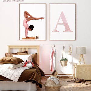 K&ouml;nnte beinhalten: Zwei gerahmte Drucke, einer mit einer Aquarellillustration einer Turnerin, die einen Handstand auf einem Schwebebalken macht, und der andere mit einer Aquarellillustration des Buchstabens A. Die Drucke h&auml;ngen an einer wei&szlig;en Wand &uuml;ber einem Holzbett mit braunen Bettw&auml;sche.