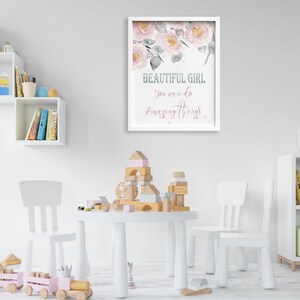 May include: A white framed print with a pink floral design and the text "BEAUTIFUL GIRL You can do Amazing Things". The print is hanging on a white wall in a child's playroom with a white table and chairs, wooden blocks, and a toy train.