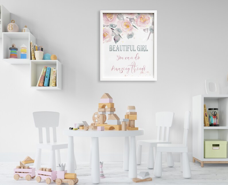 May include: A white framed print with a pink floral design and the text "BEAUTIFUL GIRL You can do Amazing Things". The print is hanging on a white wall in a child's playroom with a white table and chairs, wooden blocks, and a toy train.