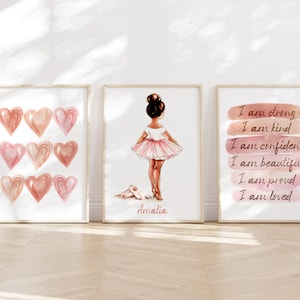 May include: Three framed prints with a pink and white color scheme. The left print features a pattern of nine pink hearts. The center print shows a ballerina in a pink tutu with the name "Amalia" written below. The right print has a watercolor background with the text "I am strong, I am kind, I am confident, I am beautiful, I am proud, I am loved."
