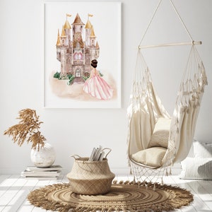 May include: A framed watercolor print featuring a large castle with golden spires and a figure in a flowing pink gown standing before it. The artwork is displayed on a white wall above a woven basket, a vase with dried grass, and a hanging chair.