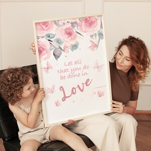 Könnte beinhalten: Ein gerahmter Kunstdruck mit einem Aquarell-Blumenmuster aus rosa Rosen und Schmetterlingen. Der Druck enthält den Text "Let all that you do be done in Love" in rosa Schrift. Der Rahmen hat eine helle Holzfarbe.