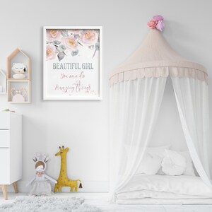 May include: A white framed print with a watercolor floral design and the text "BEAUTIFUL GIRL You can do amazing things!" hanging on a white wall. A white canopy bed with a pink canopy and white pillows is in the foreground.