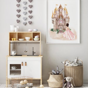May include: A wooden toy kitchen with white doors and a metal sink, featuring a framed watercolor print of a castle and a person in a pink dress above it. A hanging heart mobile, two woven baskets with plush toys, and wooden balls are also present.