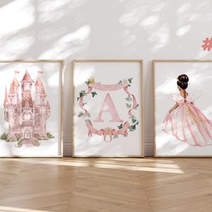 May include: Three framed watercolor prints featuring a pink castle, a pink letter A in a wreath, and a pink fairy with wings. The prints are in wooden frames and are hanging on a white wall.