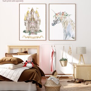 K&ouml;nnte beinhalten: Zwei gerahmte Aquarelldrucke, einer zeigt ein M&auml;rchenschloss mit goldenen T&uuml;rmen und einem Schleifenbanner, der andere zeigt ein wei&szlig;es Einhorn mit einer Blumenkrone. Die Drucke werden an einer wei&szlig;en Wand &uuml;ber einem Holzbett mit braunen Bettw&auml;sche und einer wei&szlig;en Tagesdecke angezeigt.