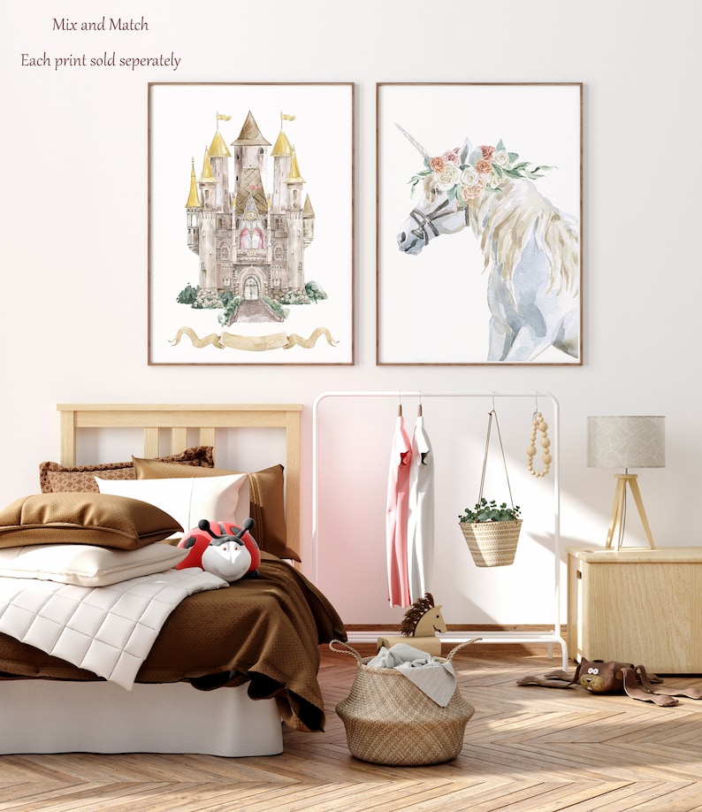 K&ouml;nnte beinhalten: Zwei gerahmte Aquarelldrucke, einer zeigt ein M&auml;rchenschloss mit goldenen T&uuml;rmen und einem Schleifenbanner, der andere zeigt ein wei&szlig;es Einhorn mit einer Blumenkrone. Die Drucke werden an einer wei&szlig;en Wand &uuml;ber einem Holzbett mit braunen Bettw&auml;sche und einer wei&szlig;en Tagesdecke angezeigt.