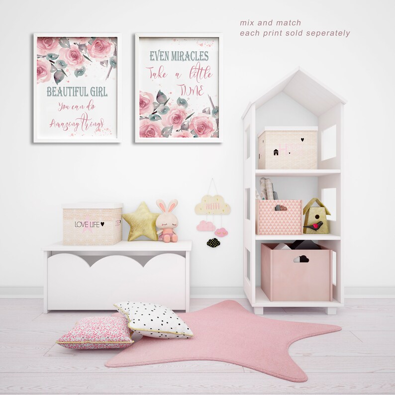 May include: Two framed watercolor prints with pink roses and inspirational quotes. The prints are hung on a white wall above a white shelf with pink and white storage boxes. The shelf is part of a white dollhouse-style bookcase.