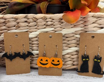 May include: Three pairs of Halloween-themed earrings displayed on brown cards. One pair features black bats, another shows orange jack-o'-lanterns, and the last depicts black cats. The earrings are set against a backdrop of autumn decor.