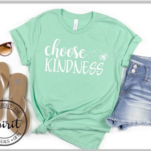 Pu&ograve; includere: Una maglietta verde chiaro con il testo "choose kindness" in lettere bianche. Un piccolo grafico di un'ape &egrave; a destra del testo. La maglietta &egrave; disposta su una superficie bianca con un paio di infradito marroni e un paio di pantaloncini di jeans blu chiaro.