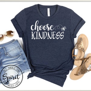 Pu&ograve; includere: Una maglietta blu navy con il testo "choose kindness" in lettere bianche. Un piccolo grafico di un'ape &egrave; sopra il testo. La maglietta &egrave; stesa su uno sfondo bianco con un paio di occhiali da sole marroni e pantaloncini di jeans.