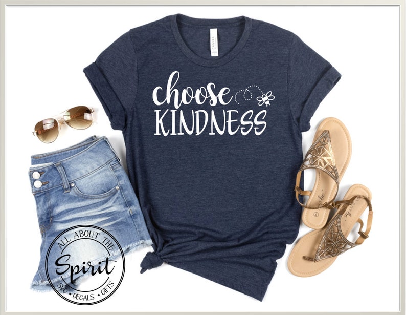 Pu&ograve; includere: Una maglietta blu navy con il testo "choose kindness" in lettere bianche. Un piccolo grafico di un'ape &egrave; sopra il testo. La maglietta &egrave; stesa su uno sfondo bianco con un paio di occhiali da sole marroni e pantaloncini di jeans.