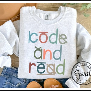 May include: A gray sweatshirt with the text "code and read" in colorful letters. The text is in a playful font and is centered on the sweatshirt. The sweatshirt is laid out on a wooden surface with a brown hat and blue jeans.